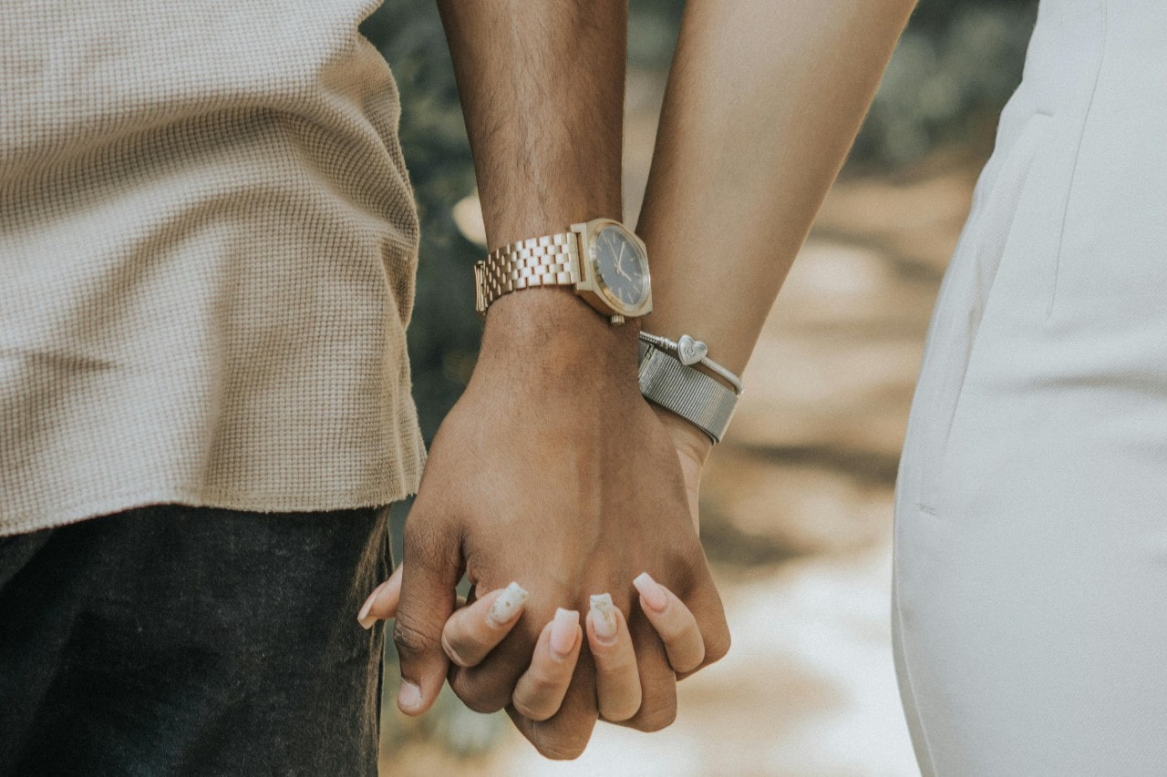 A close-up of a couple holding hands, the man wearing a yellow gold watch.