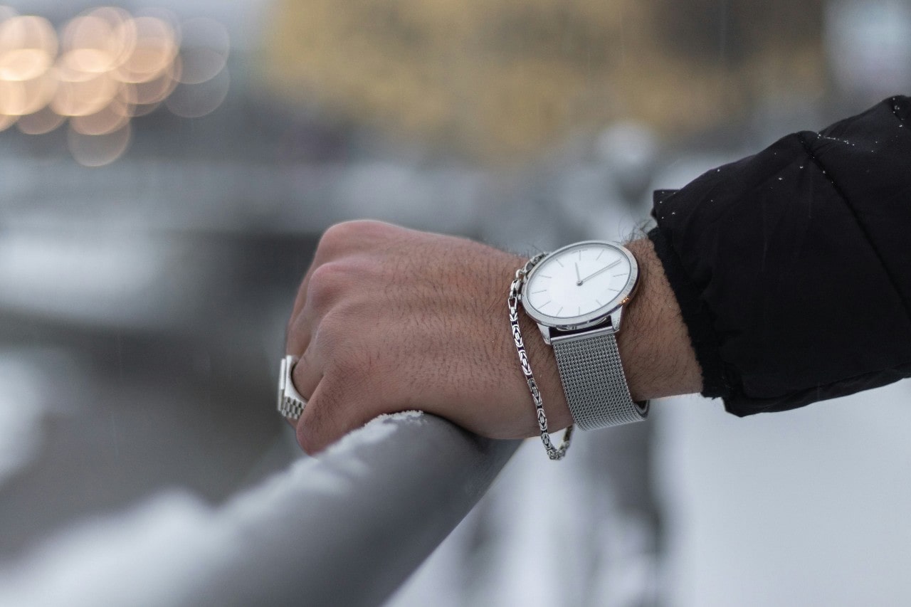 A man’s hand resting on a rail and wearing a silver minimalist watch.