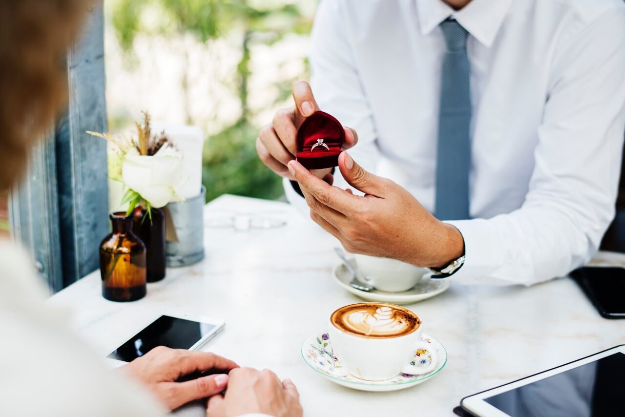 A gentleman presenting an engagement ring to his bride-to-be over coffee.