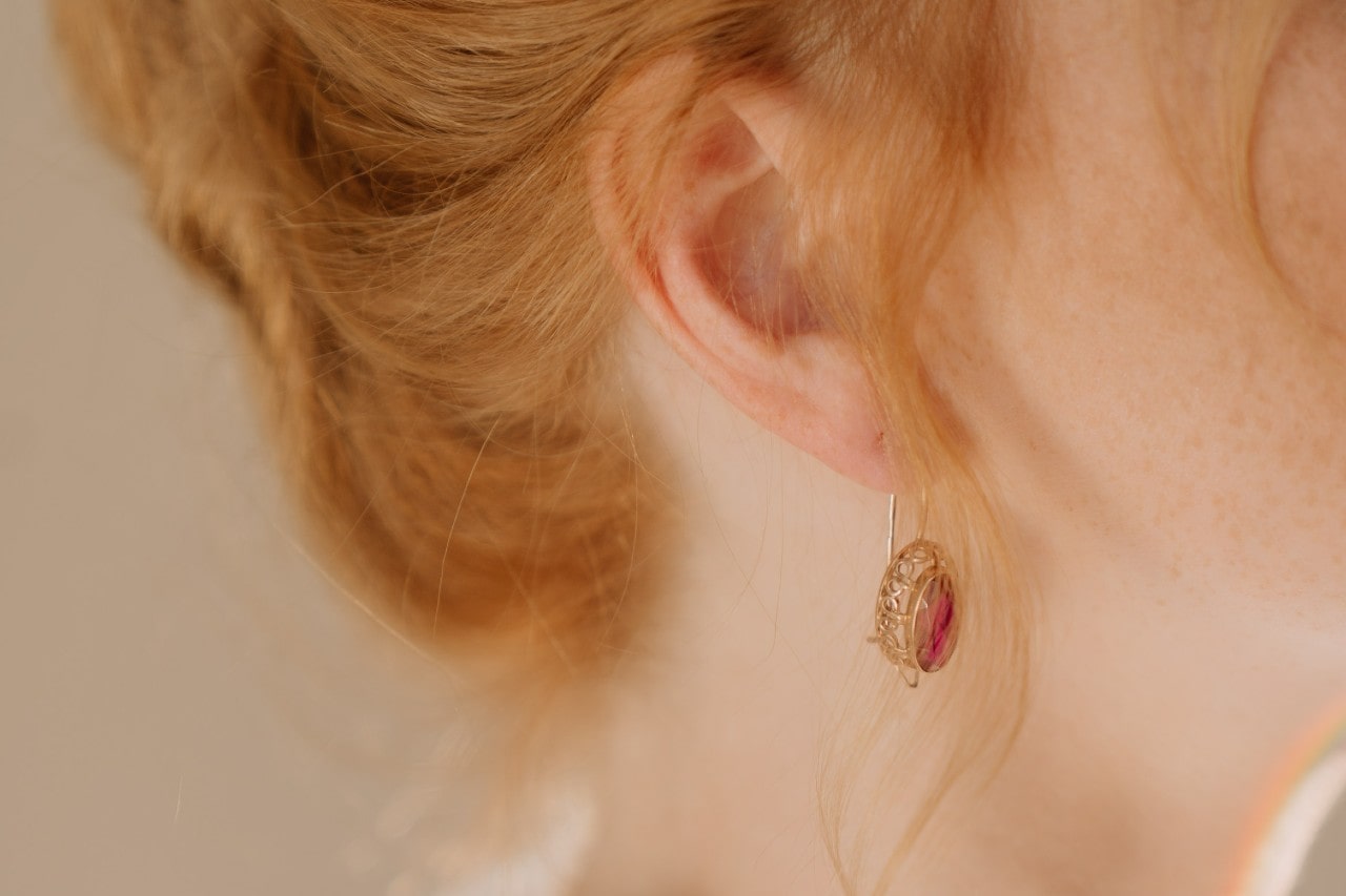 A close-up image of a woman’s ear, adorned with a yellow gold drop earring featuring a red center stone.