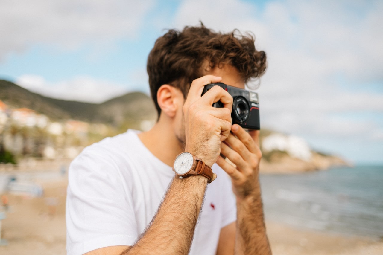 a man pointing a camera at the ocean, wearing a white and brown watch