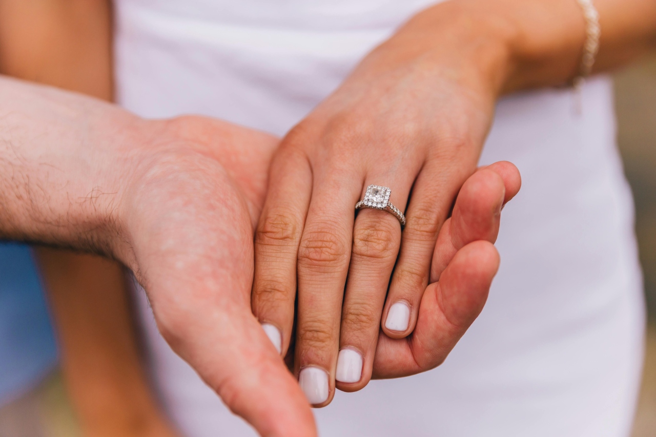 a man’s hand holding a woman’s, who is wearing a princess cut halo engagement ring