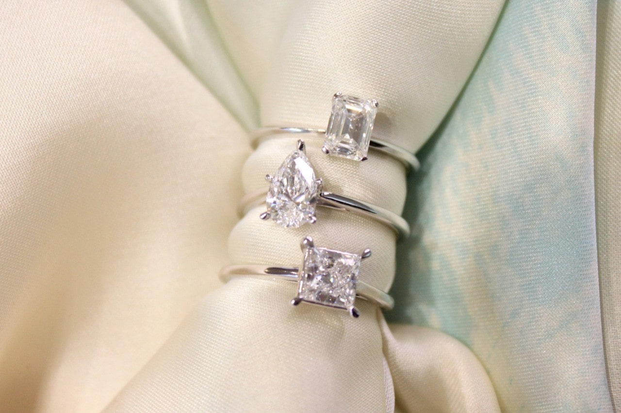 Three white gold solitaire engagement rings stacked on a piece of cream fabric.